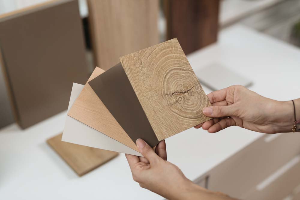 Eine Person hält Muster für Tischplatten mit verschiedenen Oberflächen, darunter Holzoptik und neutrale Farben. Im Hintergrund sind weitere größere Musterplatten sichtbar.