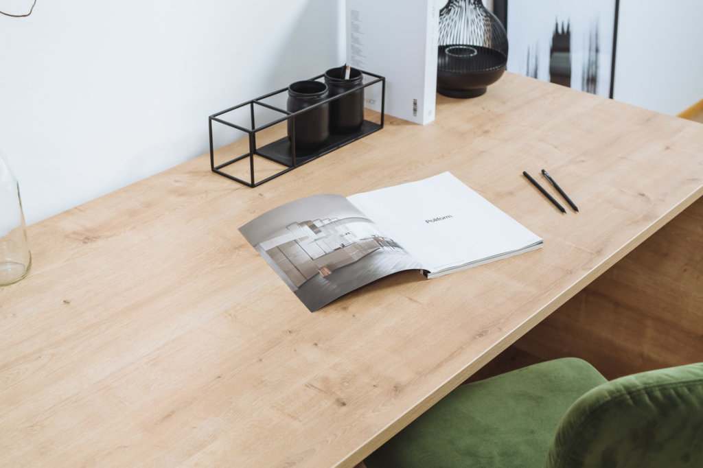 Eine Schreibtischplatte mit der Oberfläche 'Chalet Eiche' in heller Holzoptik. Darauf ein geöffnetes Magazin, zwei Stifte und ein schwarzer Organizer. Im Hintergrund eine dekorative Glasvase und weitere minimalistische Details.