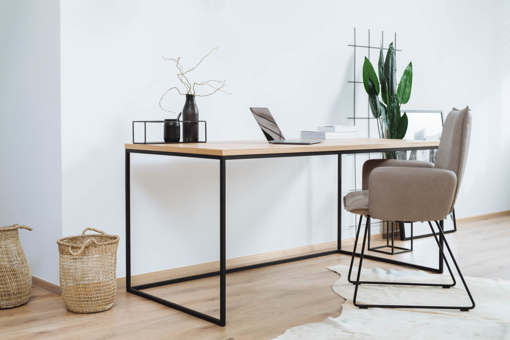 Ein minimalistischer Schreibtisch mit einer Tischplatte in Holzoptik und einem schwarzen Metallrahmen. Darauf Deko-Elemente und ein Laptop. Ergänzt wird der Raum durch einen beigen Stuhl, Pflanzen und geflochtene Körbe auf hellem Holzboden.
