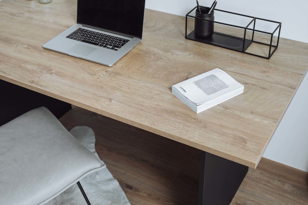 Ein Schreibtisch mit der Schreibtischplatte 'Eiche Sanremo' in natürlicher Holzoptik. Darauf liegen ein Notizbuch und ein Organizer, daneben steht ein Laptop. Die Einrichtung ist modern und minimalistisch.