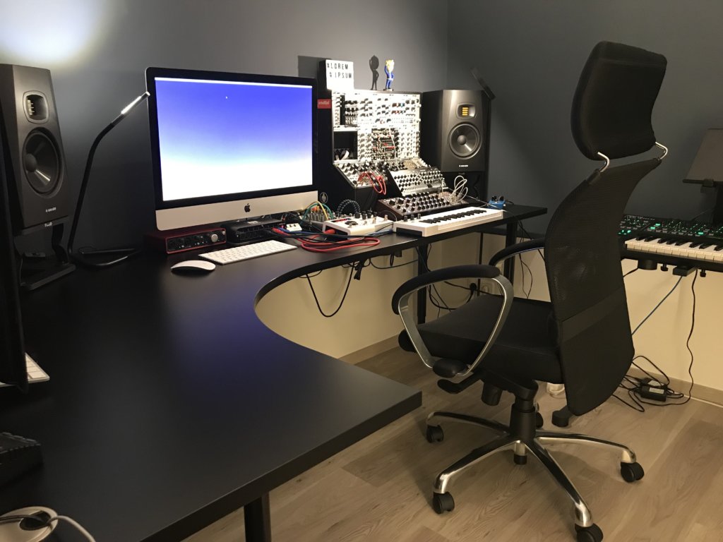 Ein ergonomischer Arbeitsplatz mit einem großen, schwarzen Eckschreibtisch, einem iMac, Studiomonitoren und modularen Synthesizern. Der Raum ist modern eingerichtet mit einer dunklen Wandfarbe und einem höhenverstellbaren Bürostuhl.