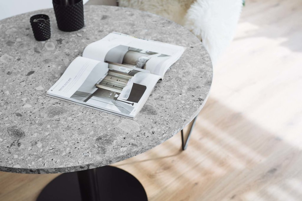 Ein runder Tisch mit einer grauen Terrazzo-Oberfläche. Darauf ein geöffnetes Magazin und dekorative schwarze Gefäße. Der Tisch steht auf einem schwarzen Metallfuß, auf hellem Holzboden.