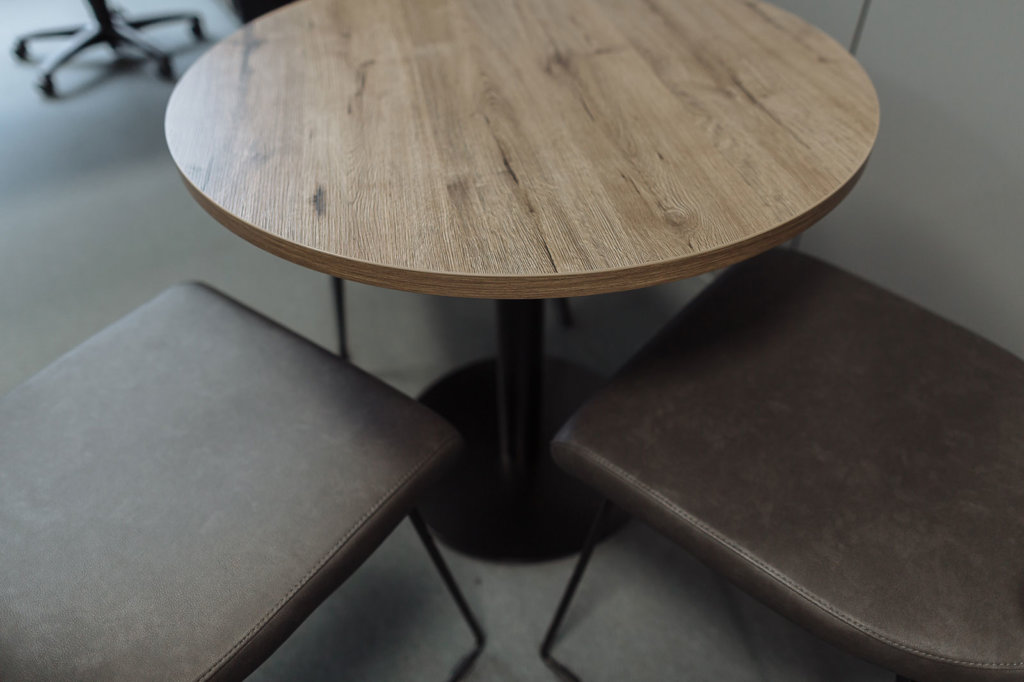 Ein runder Tisch mit einer Tischplatte in Holzoptik und einem schwarzen Metallfuß. Daneben gepolsterte Stühle mit dunkelbraunem Bezug, auf einem hellen Bodenbelag.