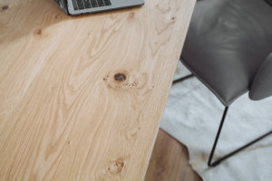 Detailaufnahme einer Tischplatte in natürlicher Holzoptik mit einer markanten Maserung. Daneben ein Stuhl mit dunkler Polsterung auf einem hellen Teppich.