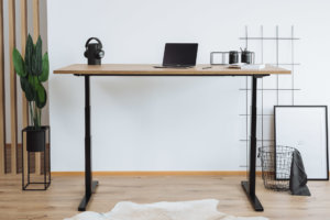 Ein höhenverstellbarer Schreibtisch mit schwarzem Gestell und einer Tischplatte in Holzoptik. Darauf ein Laptop, ein Bluetooth-Lautsprecher mit Kopfhörern sowie ein Notizbuch. Im Hintergrund eine Pflanze, ein Drahtkorb und eine minimalistische Wanddekoration.