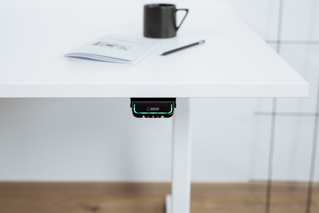Ein höhenverstellbarer Schreibtisch mit weißer Tischplatte und weißem Gestell. Darauf ein Notizbuch, ein Stift und eine Tasse, mit einem digitalen Bedienelement an der Vorderseite.