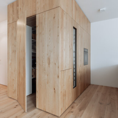 Moderne Küche mit einer Wand aus Schränken nach Maß in der Oberfläche 'Wildeiche furniert'. Die Schrankwand integriert eine Tür, die nahtlos in das Design übergeht, sowie einen Backofen und einen Weinkühlschrank. Rechts ist eine dunkle Küchenzeile mit einer Arbeitsfläche aus Stein zu sehen, während links der helle Flur mit minimalistischen weißen Türen anschließt.