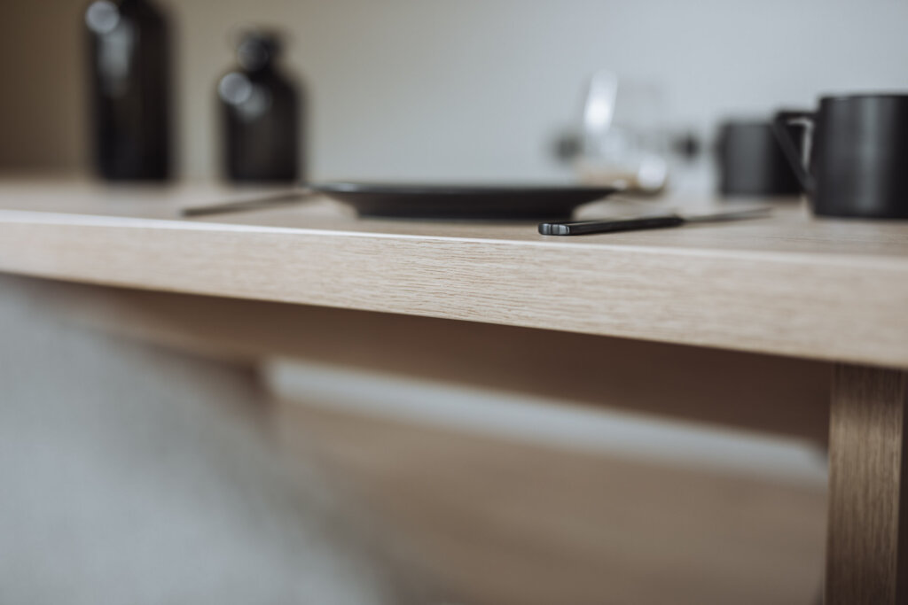 Detailaufnahme einer Tischkante aus hellem Holz mit glatter Oberfläche. Im Hintergrund befinden sich unscharf ein schwarzer Teller, Besteck und dekorative schwarze Vasen.